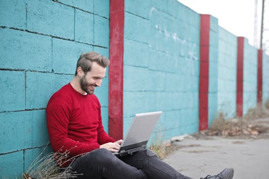 A man smiling and using a laptop outdoors against a vibrant blue wall, representing remote work.